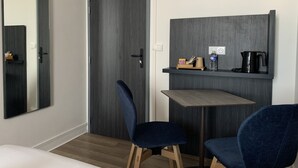 A modern room with a dark wood door, a small desk with a chair, a mirror, and a shelf with a coffee maker and a bottle. - Le Cosy Riva Bella (Ouistreham)