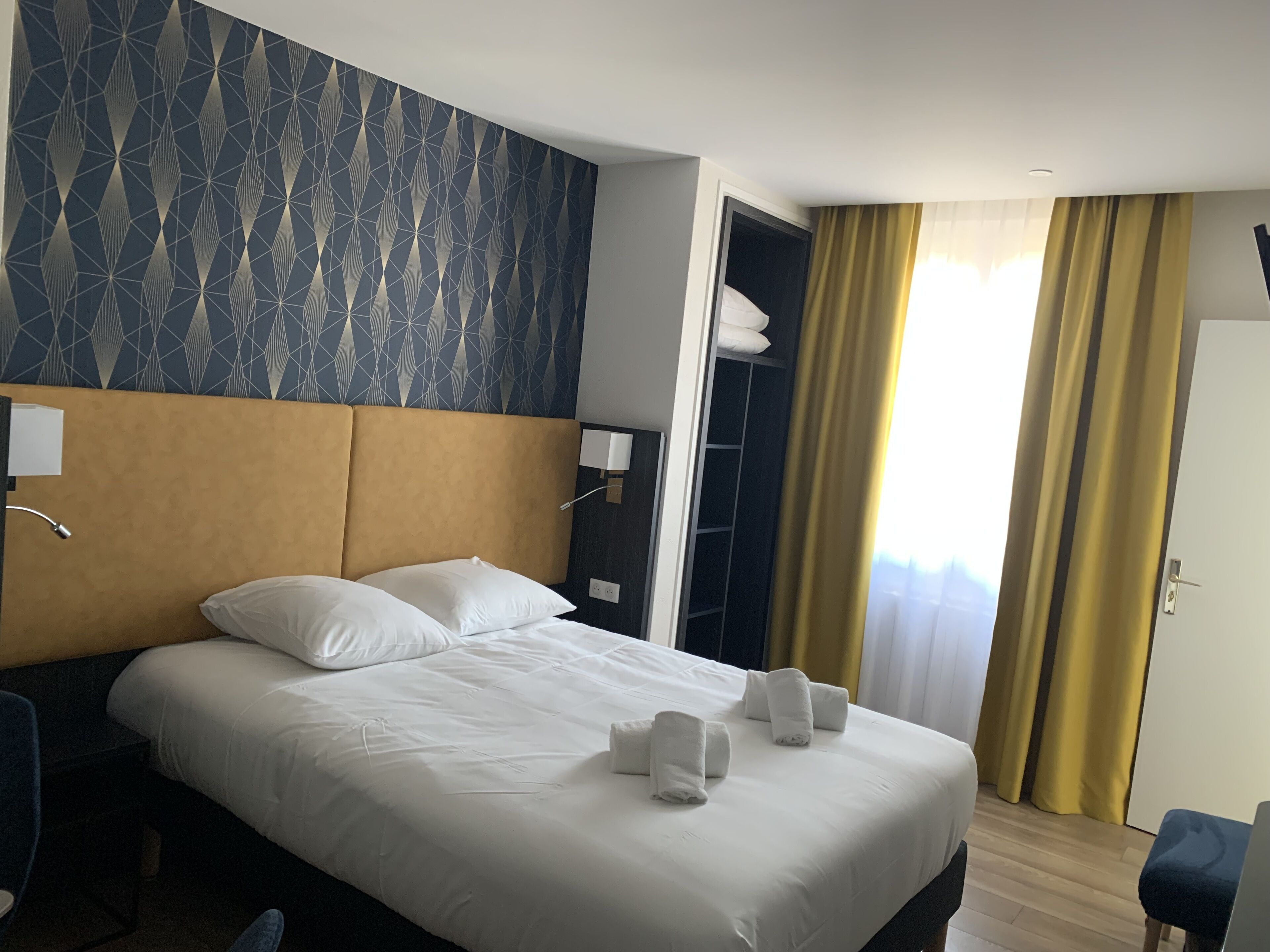 Ein Hotelzimmer mit einem großen Bett, weißen Leinentüchern, einem Kopfteil, einem Nachttisch, einem Schreibtisch, einem Sessel, einem Kleiderschrank und einem Fenster mit Vorhängen.