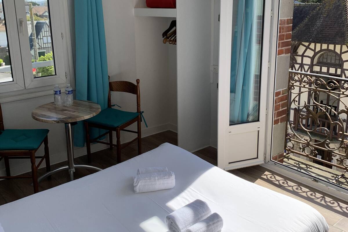 A hotel room with a bed, a small table with chairs, a window with curtains, and a balcony with a wrought iron gate.