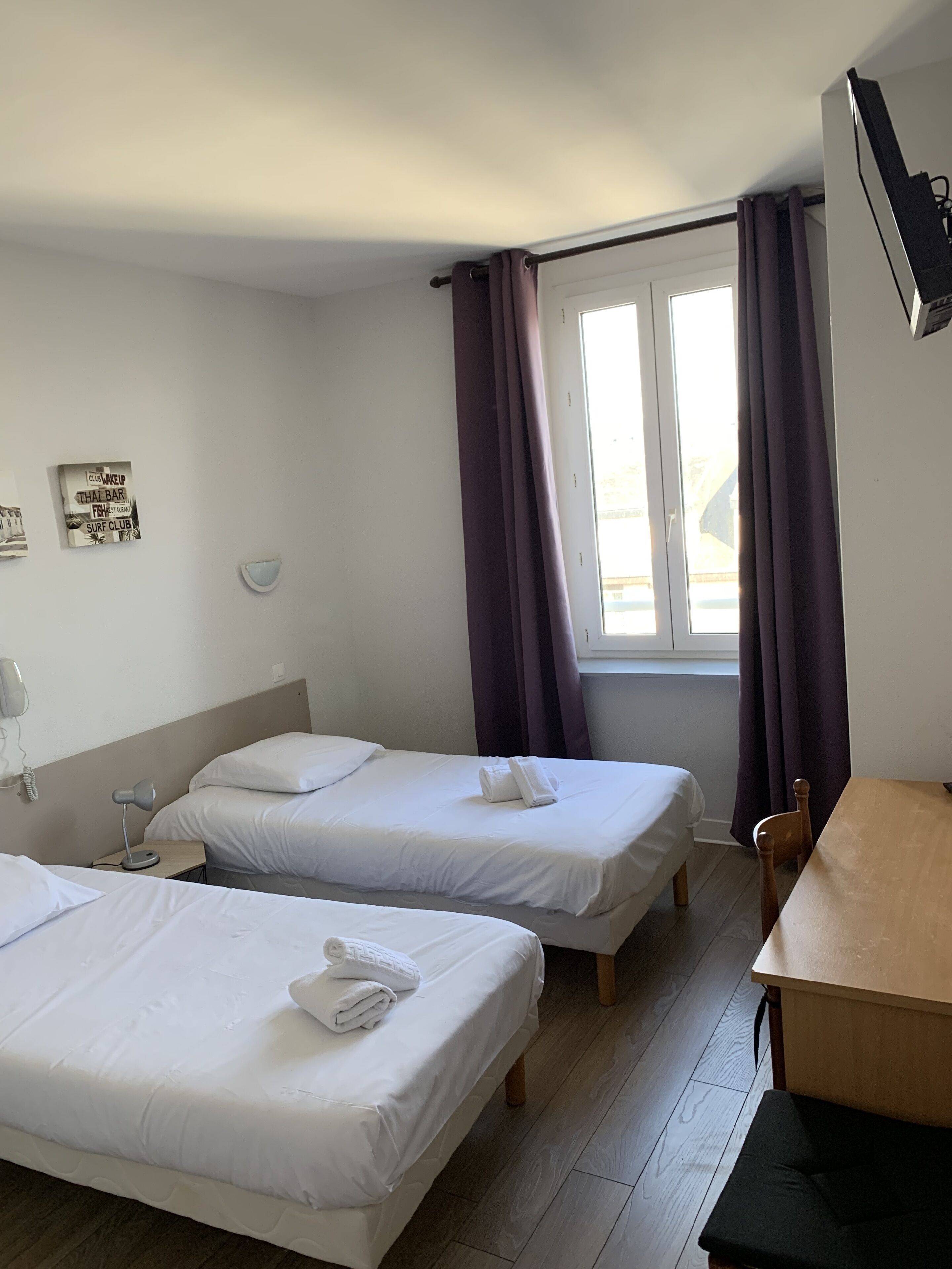 Habitación de hotel con dos camas individuales, una mesa de madera, una silla, un escritorio, una lámpara, un teléfono y una ventana con cortinas.