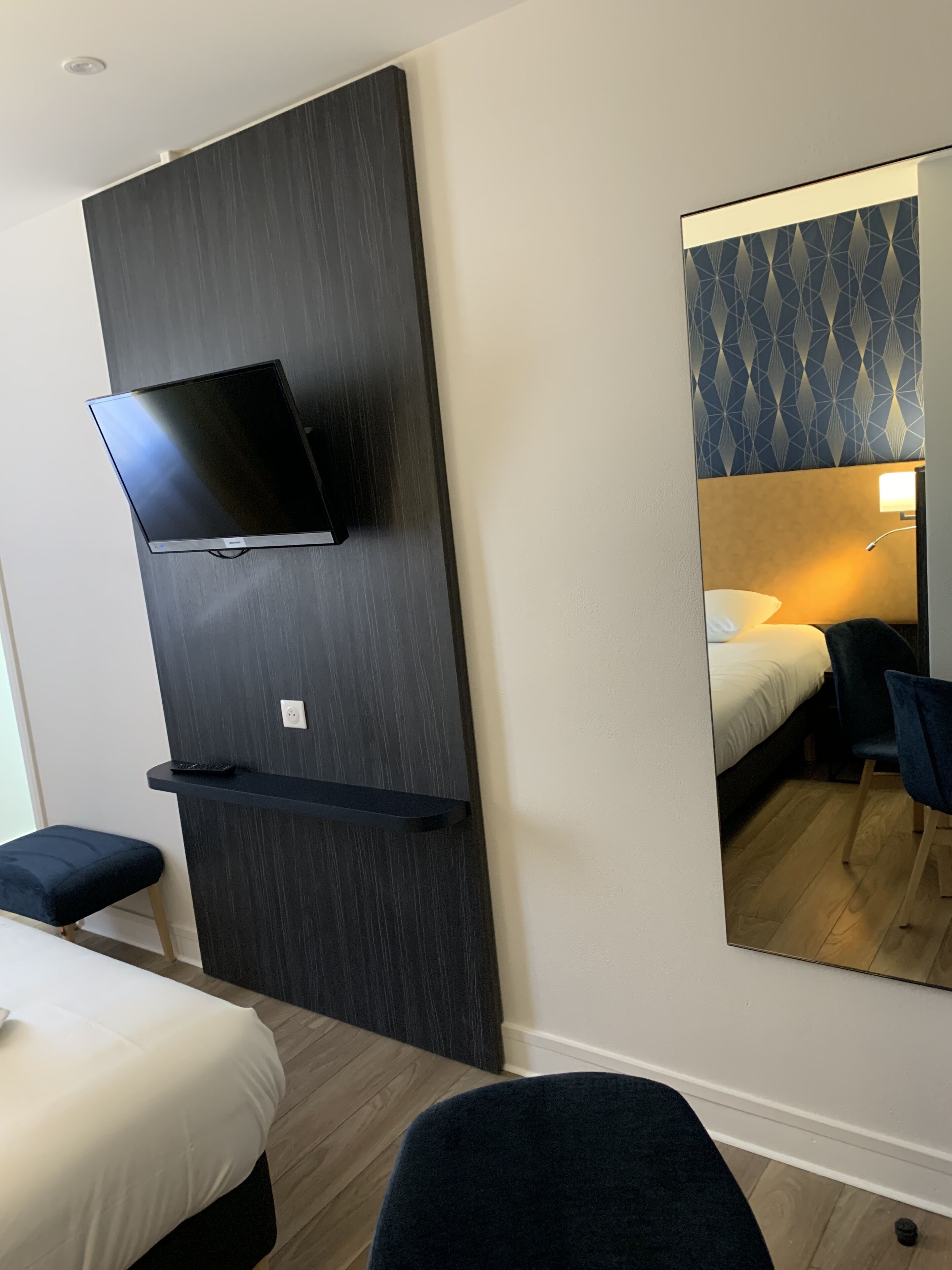 Ein Hotelzimmer mit einem Bett, einem Schreibtisch, einem Sessel, einem an der Wand befestigten Fernseher und einem Spiegel, der den Raum wiederspiegelt.