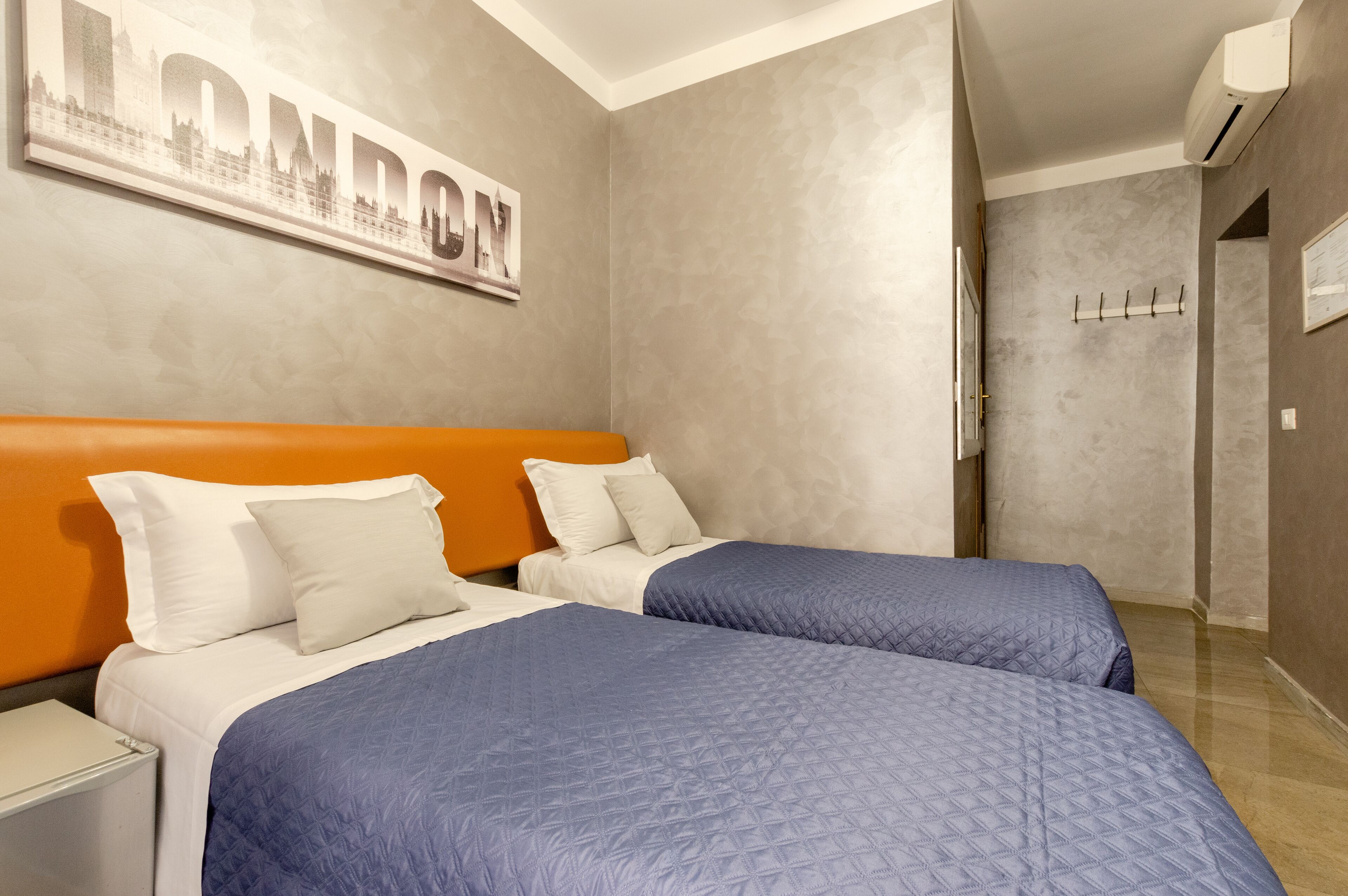 Une chambre d’hôtel avec deux lits : un grand lit à gauche et un plus petit à droite. Un tableau encadré représentant Londres est accroché au mur, au-dessus des lits.
