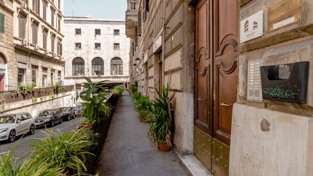 Une ruelle étroite bordée de voitures garées, de plantes en pot et d’un immeuble doté d’une grande porte en bois et de plaques commémoratives.