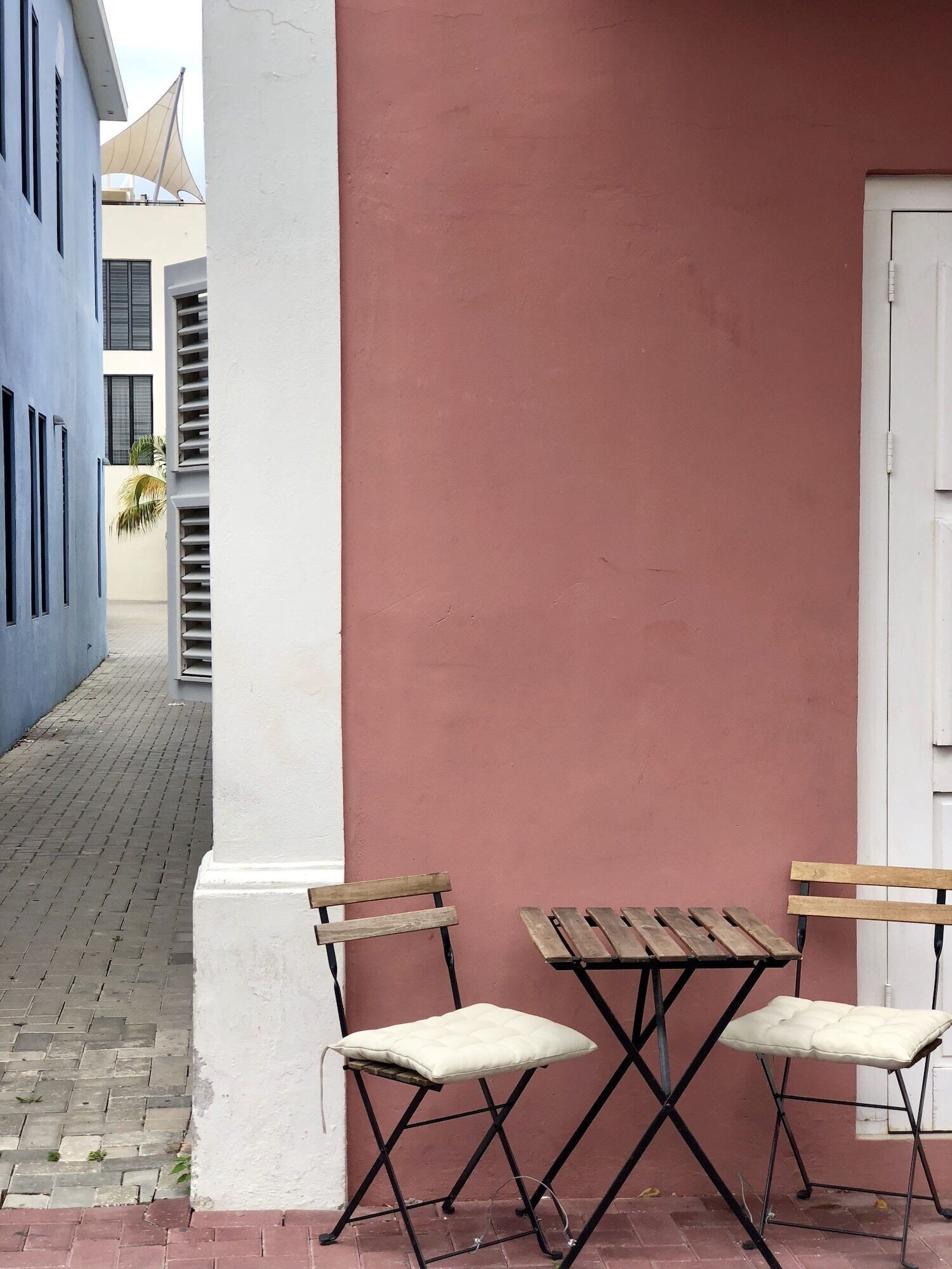 A narrow alleyway with two wooden tables and chairs, a white pillar, and a pink wall.