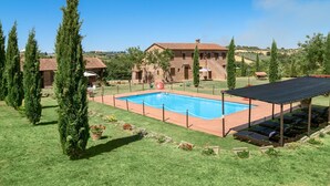 A swimming pool surrounded by a grassy area with cypress trees, a covered seating area, and a building in the background. - IL TASSO           . (Castiglione del Lago)
