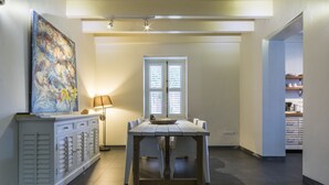 Een zithoek met een tafel, stoelen, een lamp, een schilderij en een raam met jaloezieën.