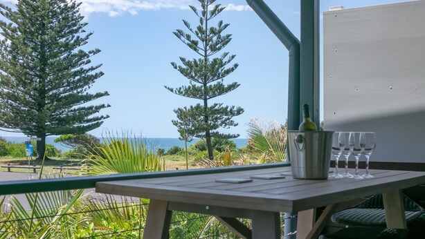 A balcony with a table, a bottle, and glasses, overlooking a coastal area with trees and the sea. - Pippis on Pacific - fantastic two-level townhouse located opposite Yamba's beautiful Pippi Beach (Yamba)