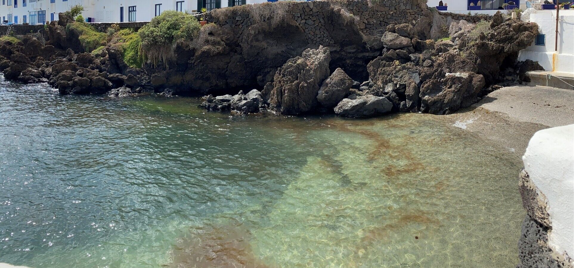 Una zona costera con aguas poco profundas y cristalinas, formaciones rocosas y un edificio blanco con ventanas azules.