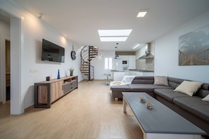 A modern living room with a sofa, coffee table, TV, wooden cabinet, spiral staircase, and a kitchen area.