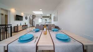 Un espace de repas moderne avec une longue table dressée pour deux personnes, un escalier en colimaçon et une cuisine en arrière-plan.