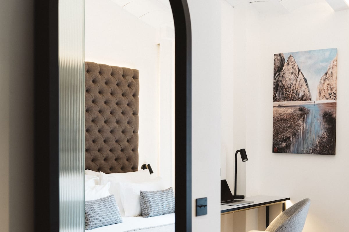 A modern bedroom with a tufted headboard, a bed with white linens, a desk with a lamp, and a chair.