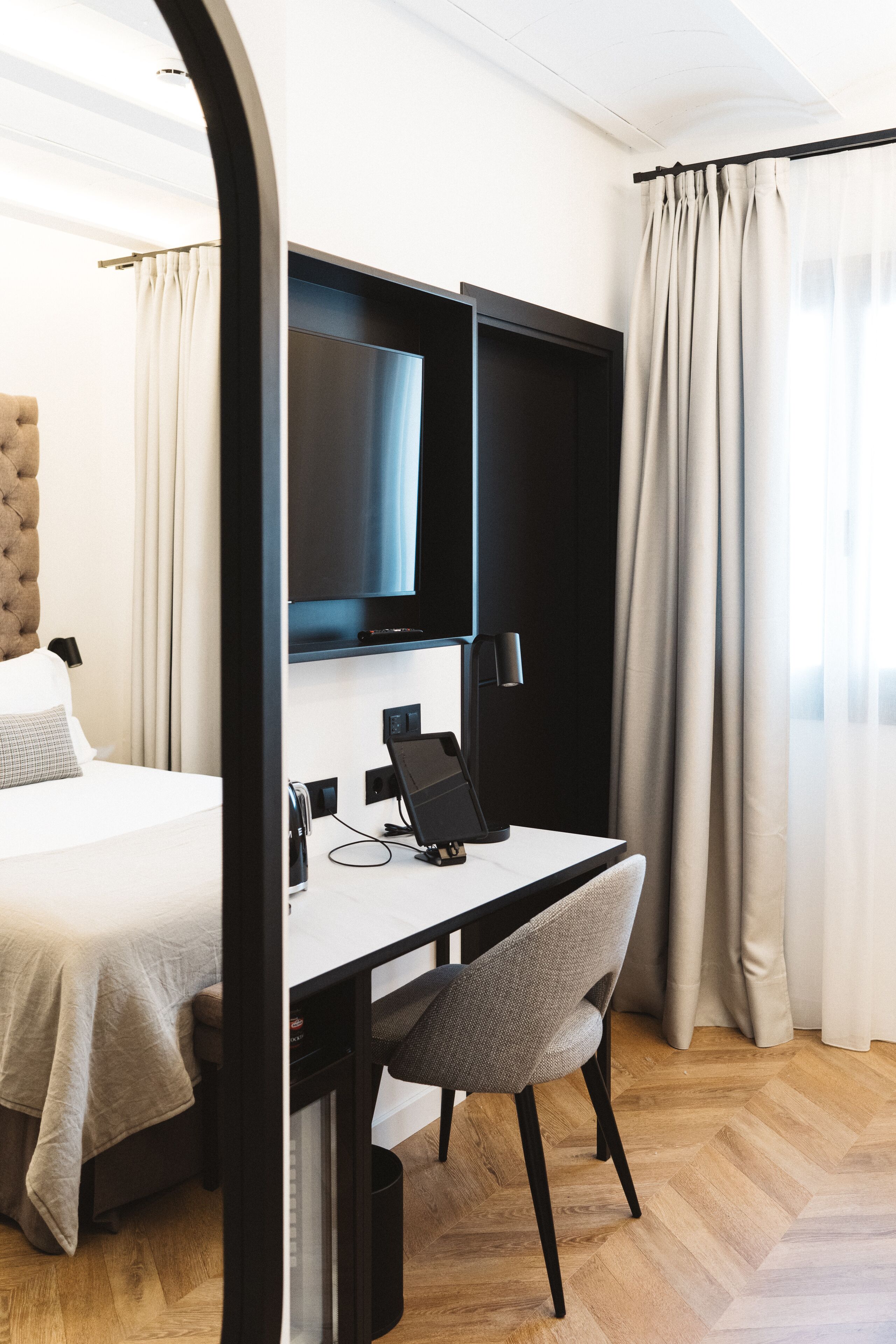 Ett hotellrum med en säng, ett skrivbord med en stol, en platt-TV och ett fönster med gardiner.