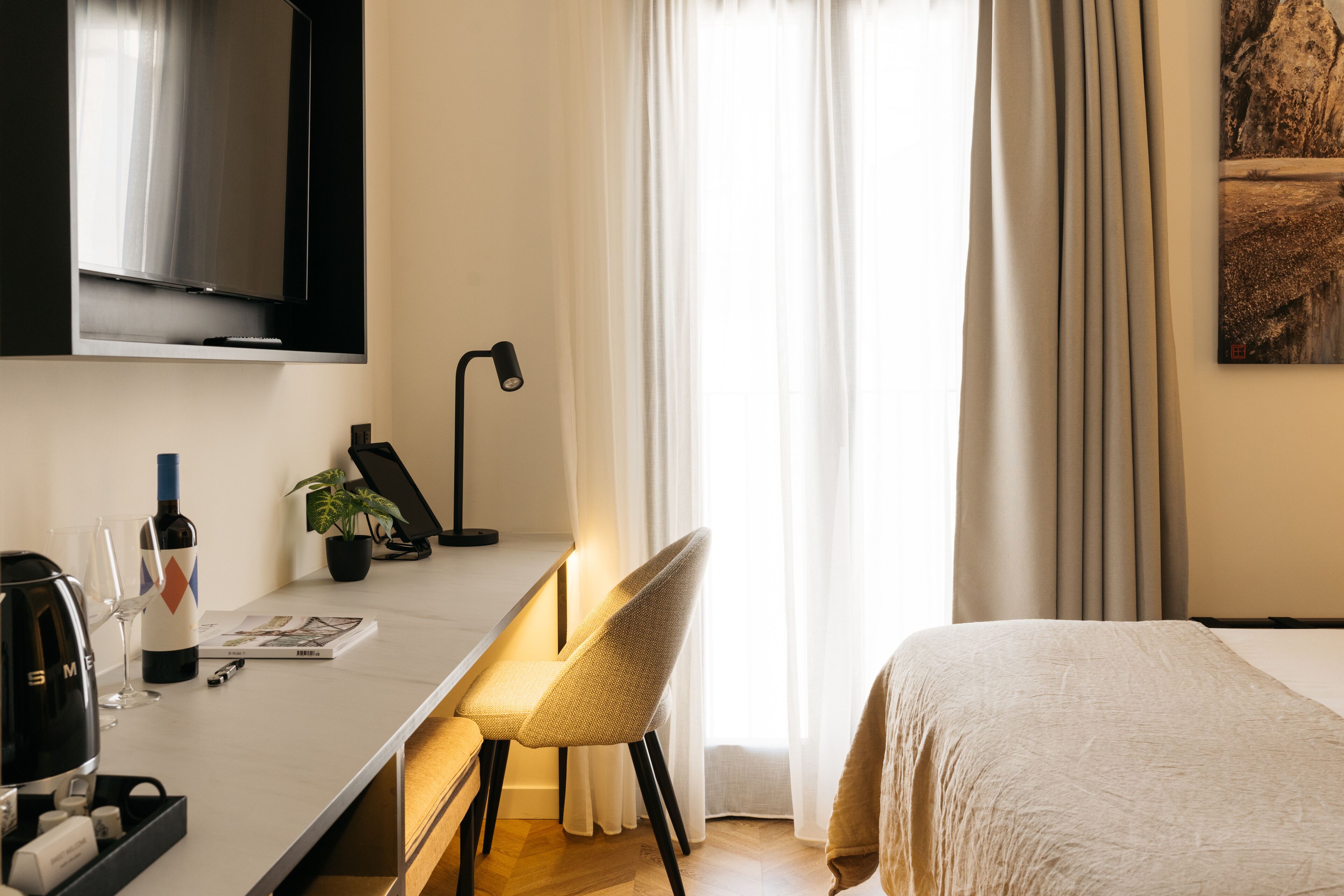 A hotel room with a bed, a desk with a chair, a television, a plant, and a window with curtains.
