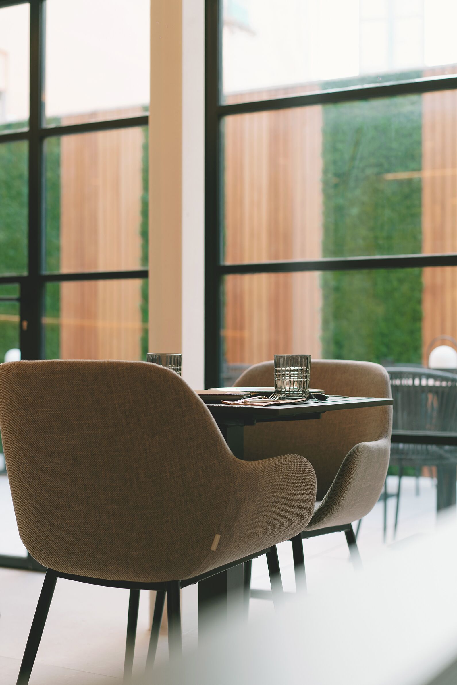 Un comedor moderno con dos sillas y una mesa, ventanas amplias y vistas a una valla de madera y follaje.