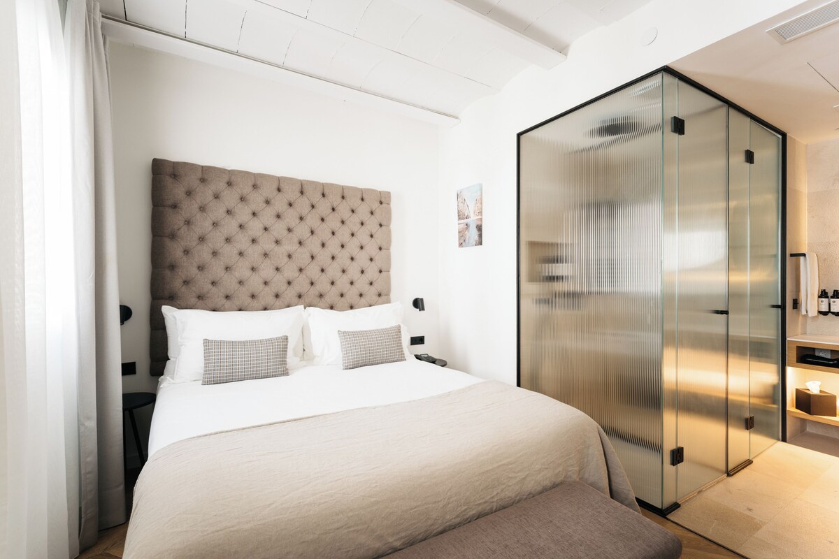Quarto moderno com cabeceira estofada, cama grande com roupa de cama branca, closet espaçoso com divisórias de vidro e mesa de cabeceira com abajur.