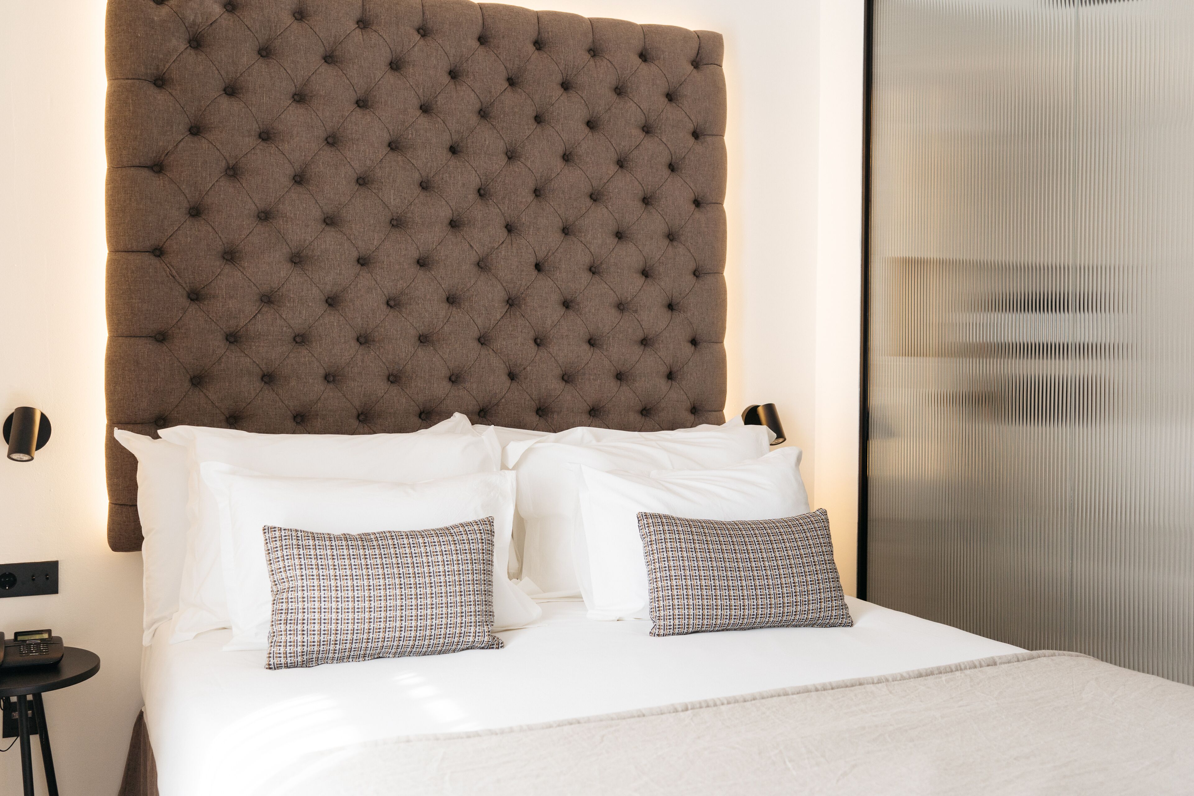 Una cama con cabecera capitonada, ropa de cama blanca y almohadas estampadas.