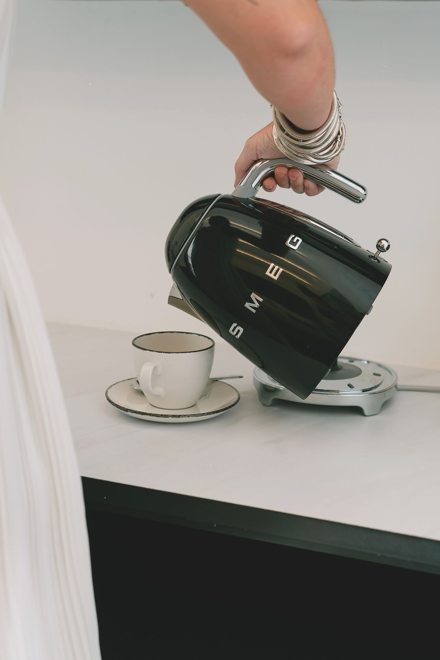 Une main tenant une bouilloire électrique noire dont on peut voir la marque, posée sur une surface blanche, à côté d’une tasse et d’une soucoupe blanches.