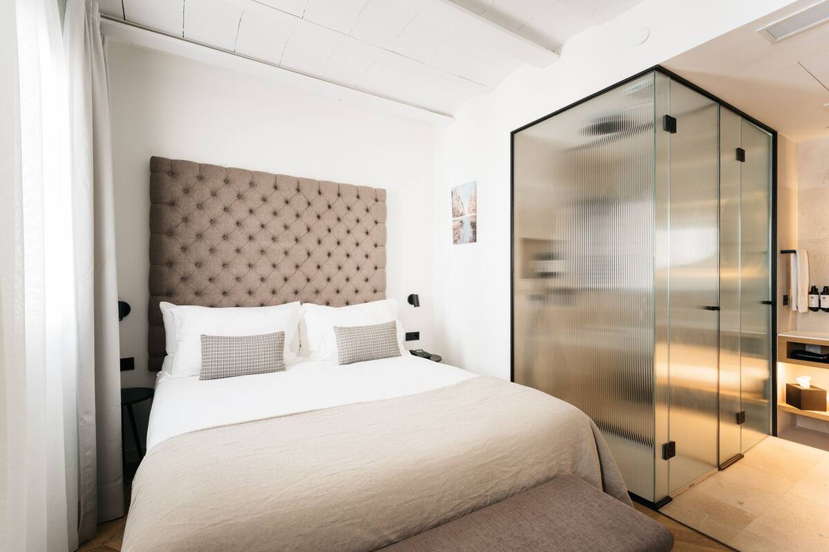 A modern bedroom with a tufted headboard, a large bed with white linens, a glass partition, and a small bedside table with a lamp.