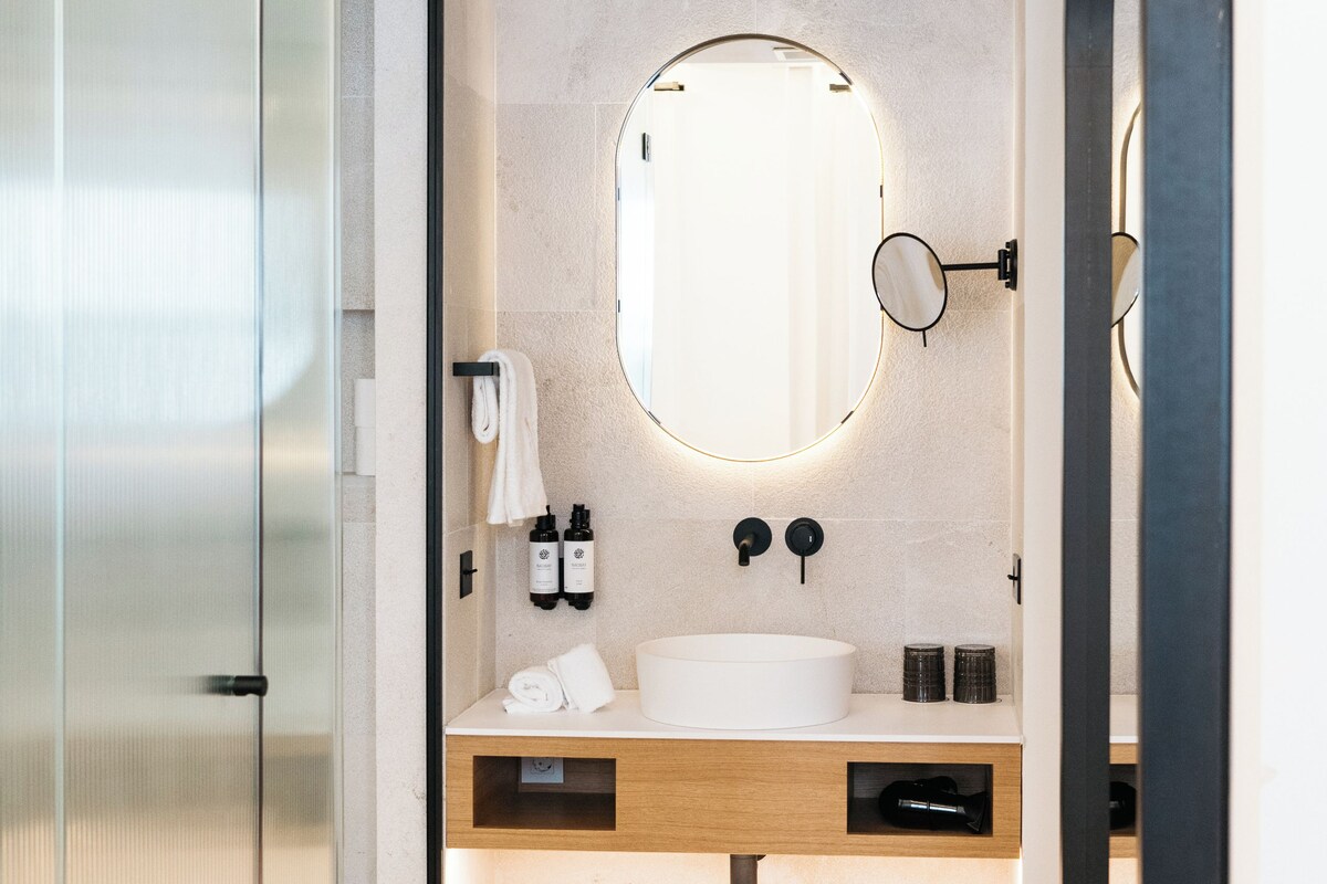 A modern bathroom with a glass shower enclosure, a round mirror, a white sink, and a wooden vanity.