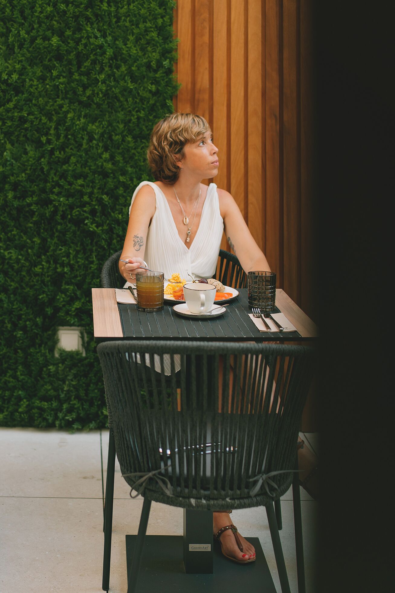 A person seated at a table with a plate of food, a glass of drink, and a cup. The table is set on a tiled floor with a green hedge in the background.