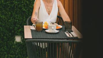 A person seated at a table with a plate of food, a glass of drink, and a cup. The table is set on a tiled floor with a green hedge in the background.