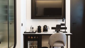 A hotel room with a flat-screen TV mounted on the wall, a small refrigerator stocked with beverages, a coffee maker, and a chair.