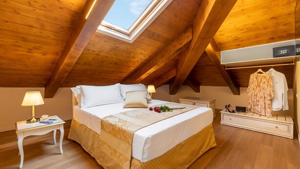 A bedroom with a skylight, wooden beams, a bed with a white and gold bedspread, a bedside table with a lamp, and a clothing rack with a dress. - Laguna D 'oro Luxury Apartments - Exclusive Sky Suite (Venezia)