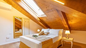 A bedroom with a sloped ceiling, a bed with white linens, a bedside table with a lamp, and a mirror on the wall. - Laguna D 'oro Luxury Apartments - Exclusive Sky Suite (Venezia)