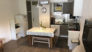 Une cuisine de taille modeste avec des meubles blancs, une table à manger recouverte d’une nappe à motifs, une horloge au mur et une petite plante en pot.