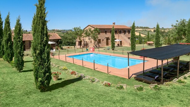 A swimming pool surrounded by a grassy area with cypress trees, a covered seating area, and a building in the background. - IL RICCIO          . (Castiglione del Lago)