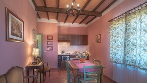 A dining area with a wooden table, chairs, and a chandelier. There is a kitchen in the background with wooden cabinets and a tiled backsplash. - IL RICCIO          . (Castiglione del Lago)