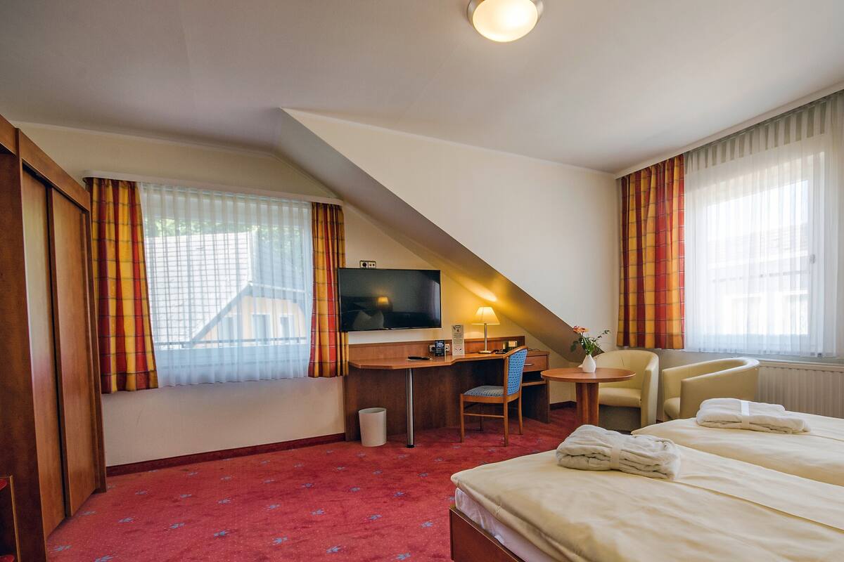 Une chambre d’hôtel avec un lit, un bureau avec un ordinateur, une chaise, une télévision et une fenêtre avec des rideaux.