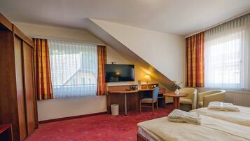 A hotel room with a bed, a desk with a computer, a chair, a TV, and a window with curtains.