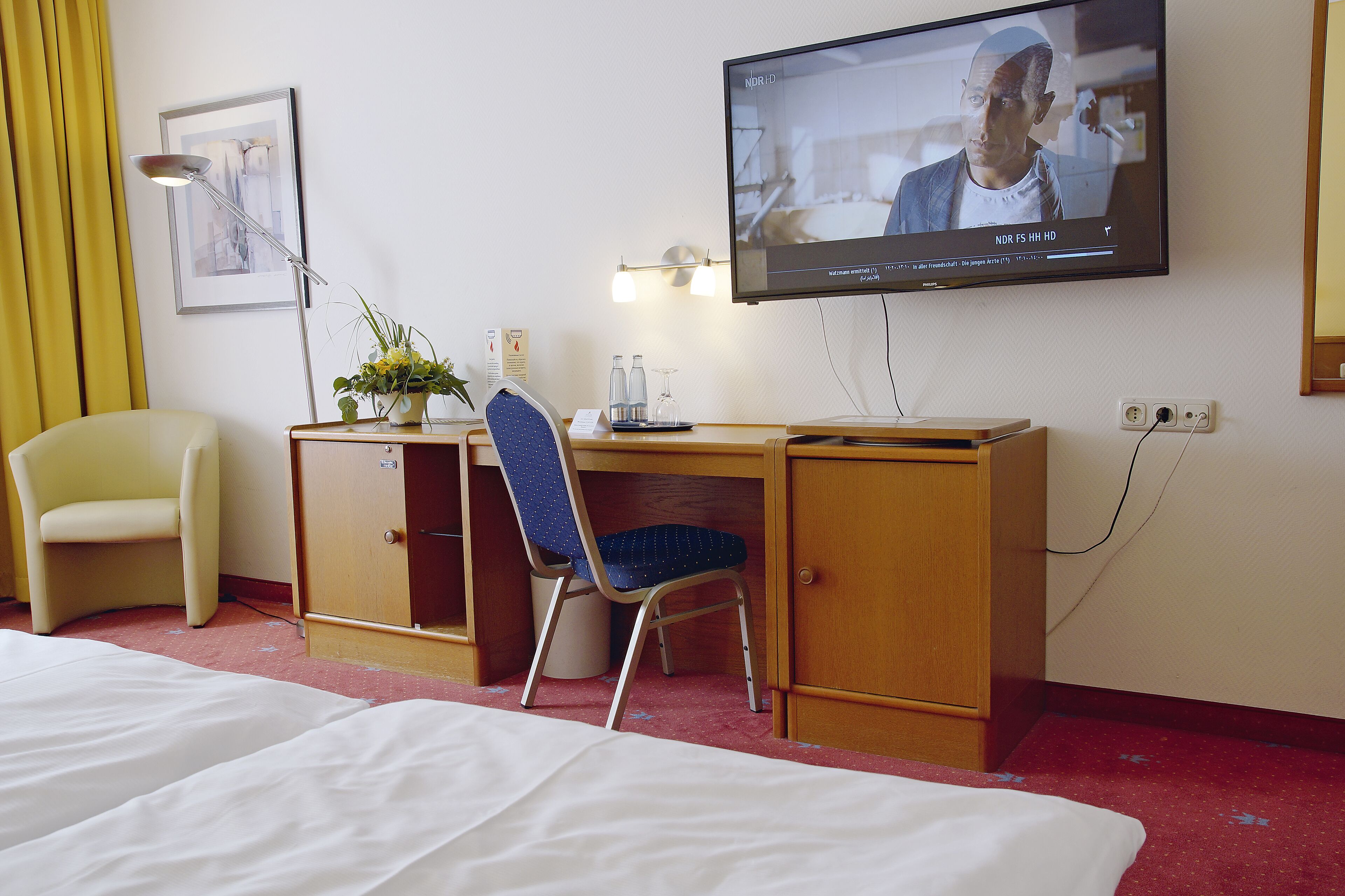 Habitación de hotel con cama, escritorio, silla, televisión y una pequeña mesa con un florero.
