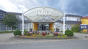 A hotel entrance with a covered entrance, a flower bed with various flowers, and a signboard. - AVALON Hotelpark Königshof (Königslutter am Elm)