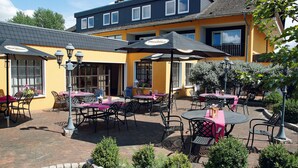 An outdoor patio area with tables and chairs, covered by umbrellas, and a building with a yellow facade in the background. - AVALON Hotelpark Königshof (Königslutter am Elm)