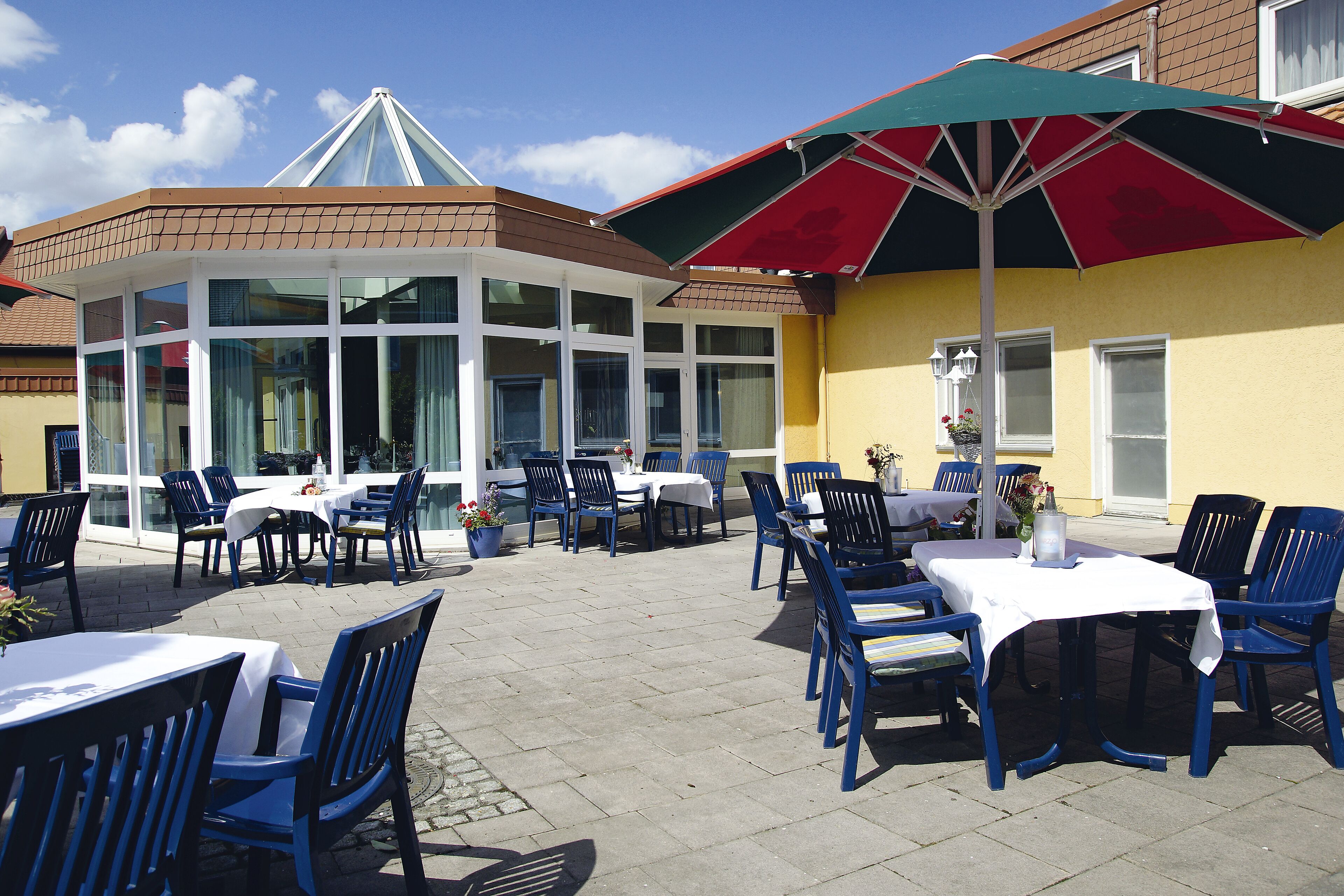 Un patio al aire libre con mesas y sillas, una zona de estar cubierta y un edificio con ventanas amplias.
