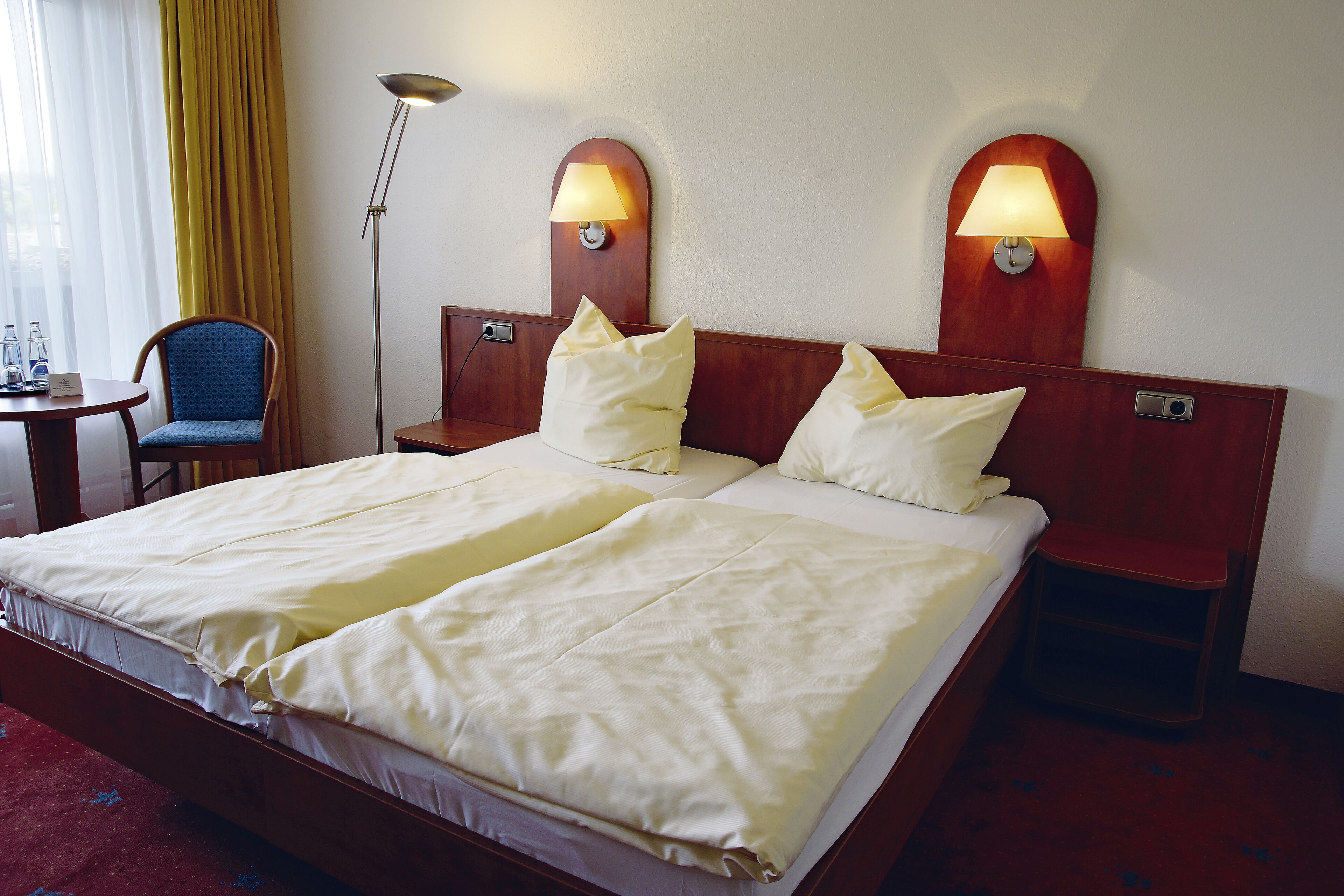 A hotel room with a double bed, two bedside tables, a chair, a lamp, and a window with curtains.