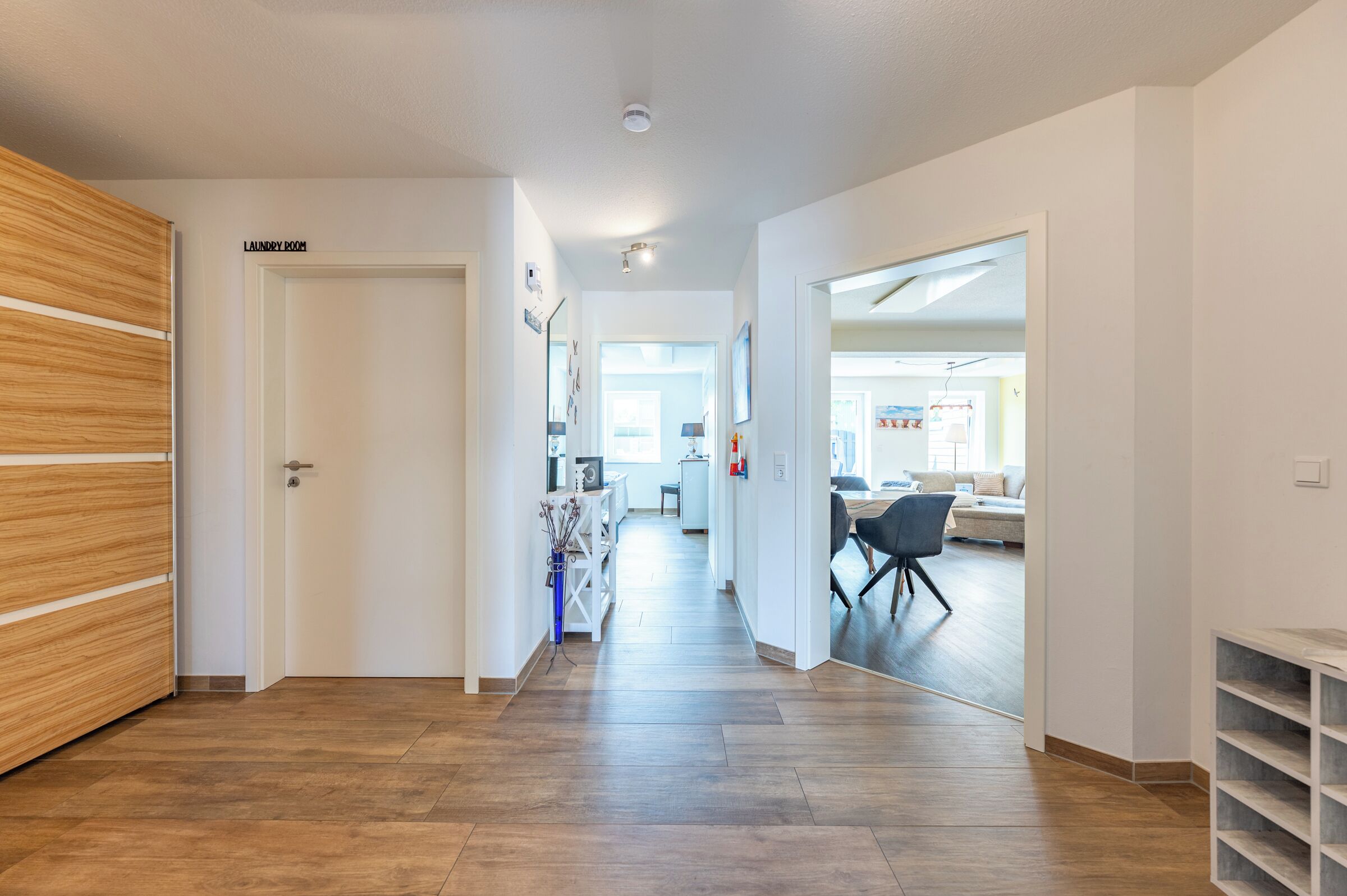 Un couloir avec une porte en bois portant l’inscription « buanderie », un mur blanc et, au fond, un espace cuisine moderne.