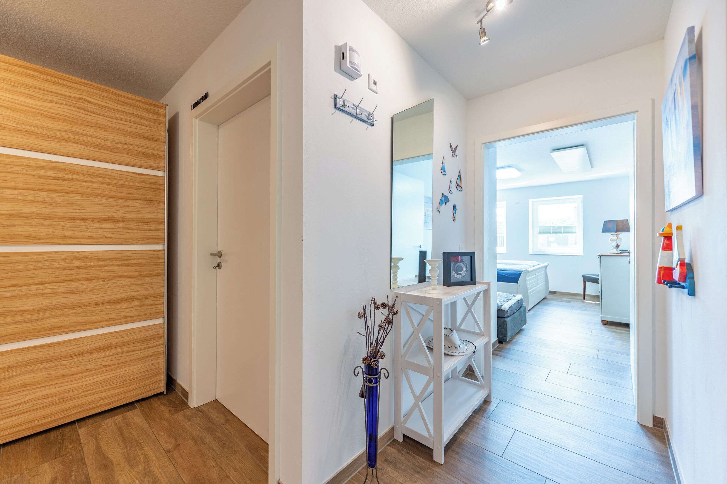Un couloir avec une porte en bois, un mur blanc orné d’un miroir et de décorations murales, et un lit à l’arrière-plan.