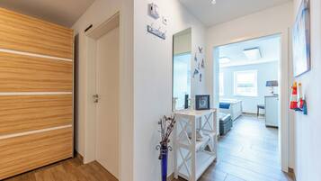 Un couloir avec une porte en bois, un mur blanc orné d’un miroir et de décorations murales, et un lit à l’arrière-plan.