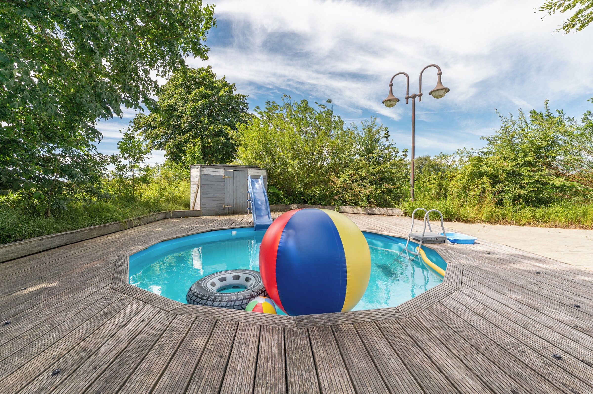 A wooden deck surrounding a swimming pool with a slide, a beach ball, and a small shed.