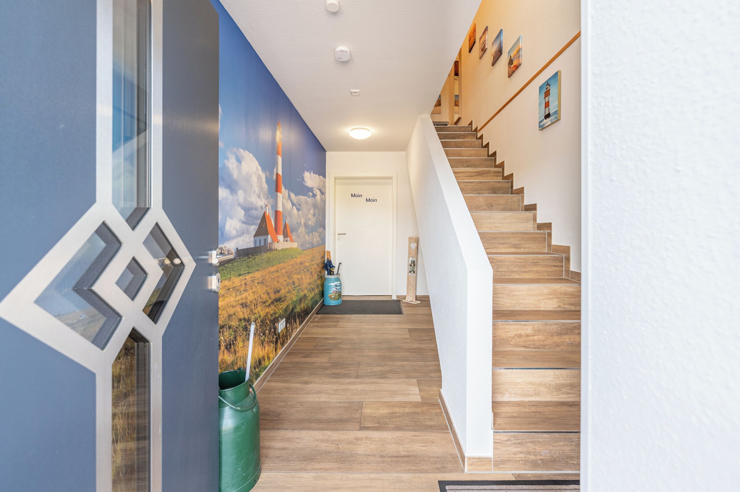 A modern interior with a wooden staircase, a mural of a lighthouse, and a door labeled 'Moin'.