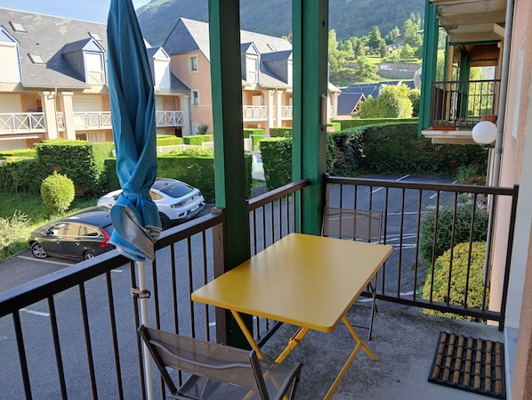A balcony with a yellow table and chairs, a blue umbrella, and a view of houses and greenery.