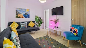 En moderne stue med en sort sofa, farverige puder, en blå lænestol, et lille rundt bord, en potteplante og et maleri på væggen.