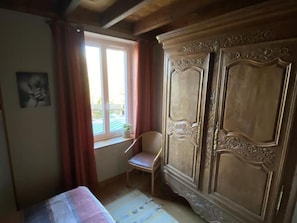 A room with a bed, a chair, a large wardrobe, a window with curtains, and a small plant on the windowsill.