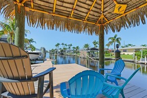 A covered deck with chairs, a thatched roof, and a marina in the background. - Two homes that exude luxury and comfort, Heated Pools - Villa Blue Pavilion- Roelens Vacations (Cape Coral)