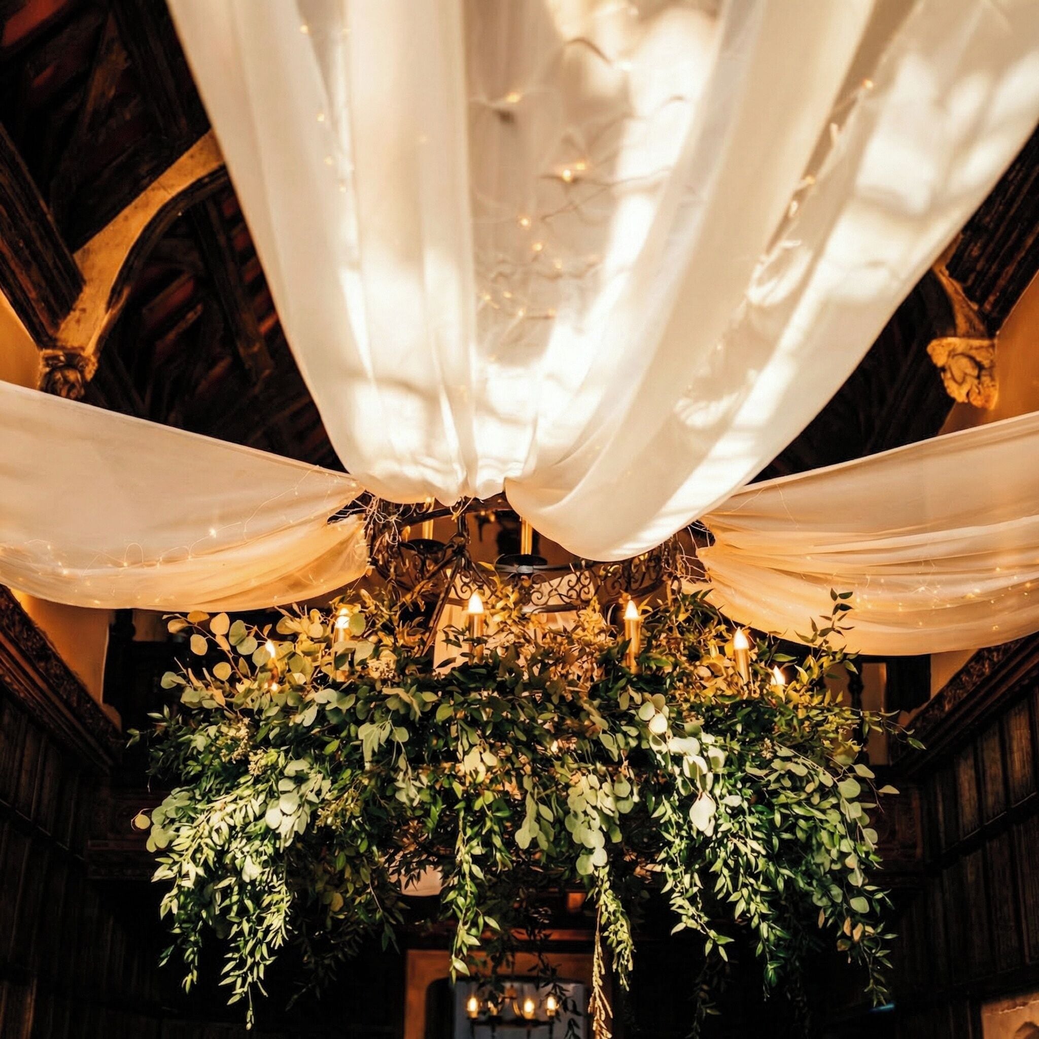 A chandelier with greenery and candles, draped fabric, and wooden beams.