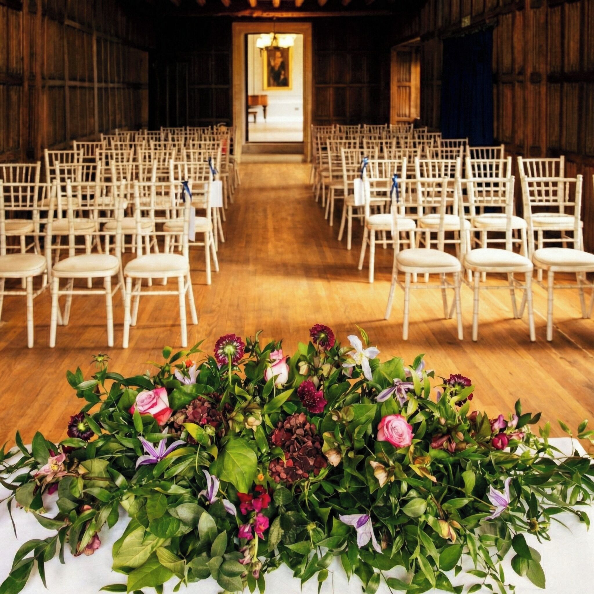 Une salle où des rangées de chaises blanches sont disposées pour un événement, un arrangement floral se trouve à l’avant, et on aperçoit au loin une autre pièce avec un lustre.