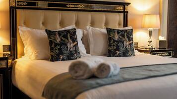 Habitación de hotel con una cama grande, dos almohadones decorativos, una lámpara de noche y una toalla enrollada sobre la cama.